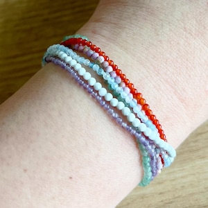 May include: A stack of seven bracelets made of different colored beads. The bracelets are made of red, white, light blue, purple, and green beads.