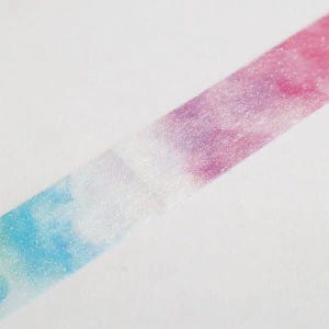 May include: A strip of adhesive tape featuring a gradient pattern that transitions from light blue to white and then to a vibrant pink and purple blend. The surface of the tape has a subtle, shimmering, glittery texture throughout its length.