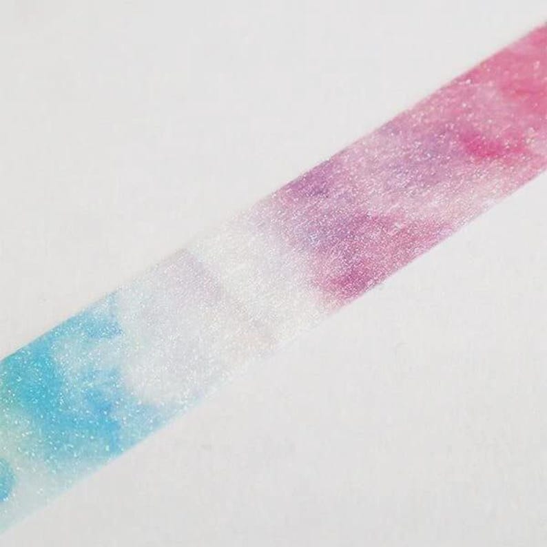 May include: A strip of adhesive tape featuring a gradient pattern that transitions from light blue to white and then to a vibrant pink and purple blend. The surface of the tape has a subtle, shimmering, glittery texture throughout its length.