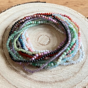 Puede incluir: Una colección de pulseras de piedras preciosas de colores apiladas sobre una superficie de madera. Las pulseras están hechas de pequeñas cuentas redondas en varios tonos de verde, azul, rosa, rojo y negro.