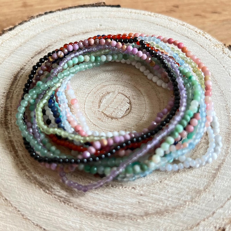 May include: A collection of colorful gemstone bracelets stacked on a wooden surface. The bracelets are made of small, round beads in various shades of green, blue, pink, red, and black.