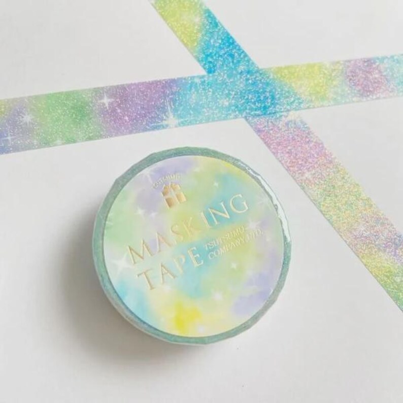 May include: A roll of masking tape with a pastel watercolor gradient of blue, green, and yellow featuring a glittery finish and white star accents. The label reads MASKING TAPE TSUTSUMU COMPANY. A strip of the same tape is laid out in an X shape.