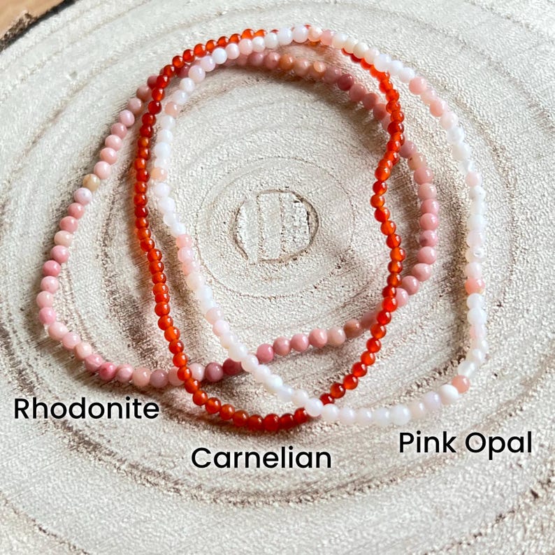May include: Three strands of gemstone bracelets, one red, one pink, and one white. The red bracelet is labeled "Carnelian", the pink bracelet is labeled "Pink Opal", and the white bracelet is labeled "Rhodonite".