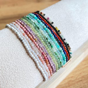 May include: A collection of 20 colorful gemstone bracelets, each featuring a different type of stone. The bracelets are stacked on top of each other, showcasing the variety of colors and textures.