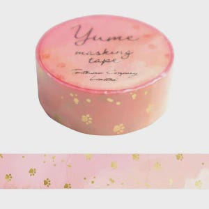 May include: A roll of pink masking tape featuring a repeating pattern of small gold paw prints. The top of the roll displays the text Yume masking tape and Tautsumi Company Limited in a dark script font against a light pink background.