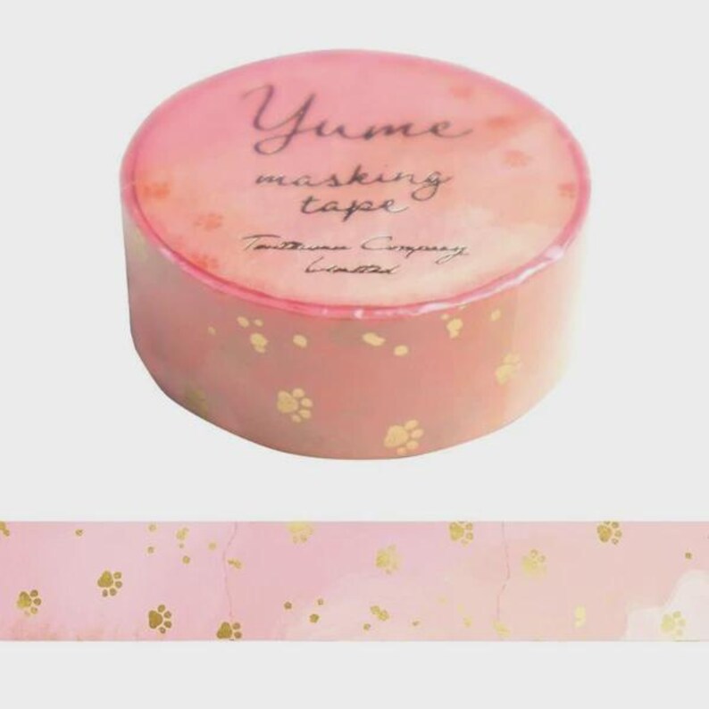 May include: A roll of pink masking tape featuring a repeating pattern of small gold paw prints. The top of the roll displays the text Yume masking tape and Tautsumi Company Limited in a dark script font against a light pink background.