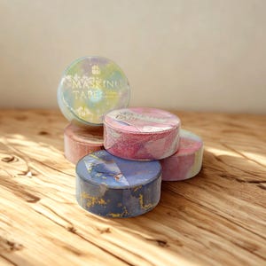 May include: A collection of five circular rolls of masking tape featuring various abstract patterns in shades of blue, pink, and pastel yellow. Each roll is labeled with the text MASKING TAPE in a metallic font, showcasing distinct marbled and watercolor-inspired designs.