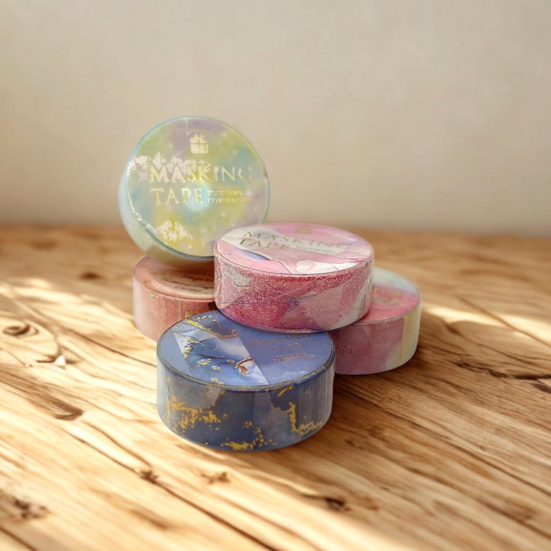 May include: A collection of five circular rolls of masking tape featuring various abstract patterns in shades of blue, pink, and pastel yellow. Each roll is labeled with the text MASKING TAPE in a metallic font, showcasing distinct marbled and watercolor-inspired designs.