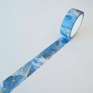 May include: A roll of washi tape featuring a blue and white watercolor-style marble pattern with metallic gold foil accents. The tape is partially unrolled, displaying a continuous abstract design with flowing blue tones and gold flecks.
