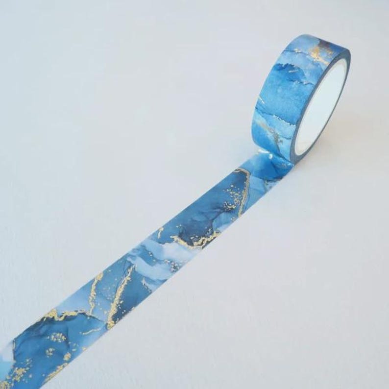 May include: A roll of washi tape featuring a blue and white watercolor-style marble pattern with metallic gold foil accents. The tape is partially unrolled, displaying a continuous abstract design with flowing blue tones and gold flecks.