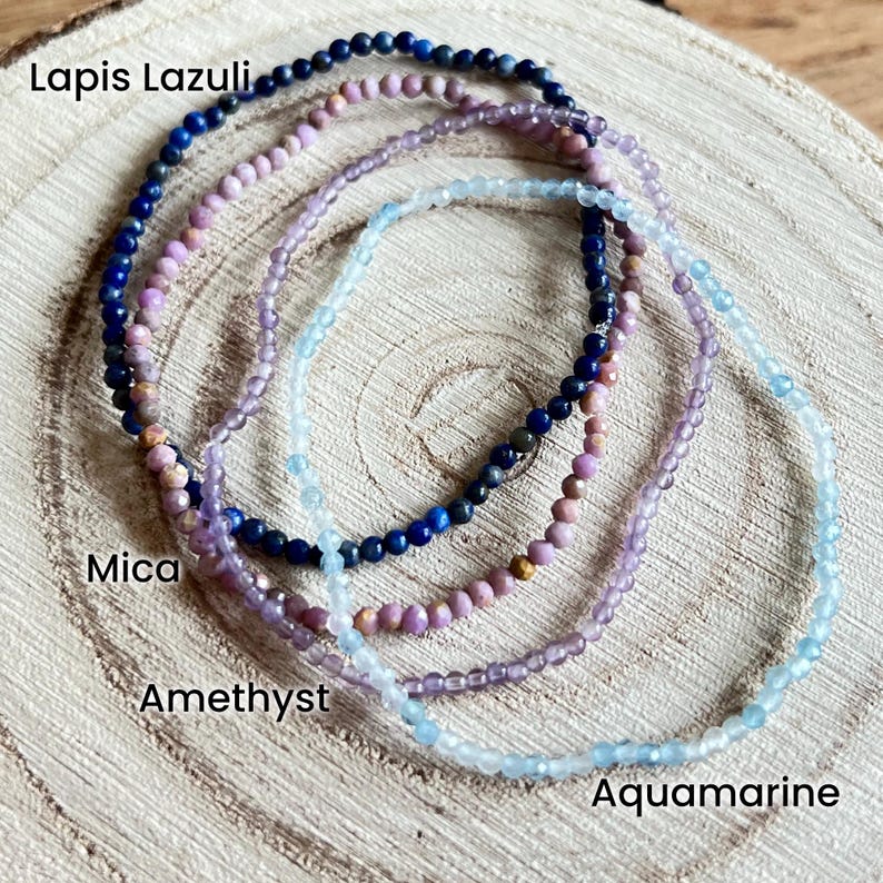 May include: Four gemstone bracelets stacked on a wooden surface. The bracelets are made of lapis lazuli, mica, amethyst, and aquamarine beads. The bracelets are labeled with their respective gemstone names.