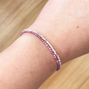 May include: A delicate bracelet with two strands of small, colorful beads. The top strand is made of white and cream beads, while the bottom strand is made of pink and purple beads.