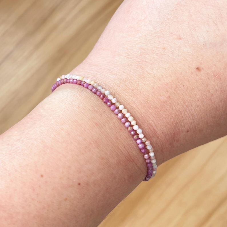 May include: A delicate bracelet with two strands of small, colorful beads. The top strand is made of white and cream beads, while the bottom strand is made of pink and purple beads.