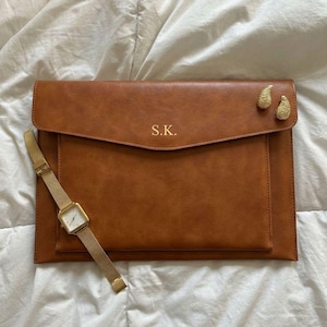 Puede incluir: Una funda de cuero marrón para portátil con las iniciales "S.K." en letras doradas. Un reloj dorado con esfera cuadrada y correa de malla dorada está junto a la funda. Dos adornos dorados decorativos están en la esquina superior derecha.
