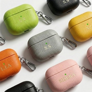 May include: A collection of colorful, textured leather AirPod cases in various colors, including green, orange, gray, yellow, pink, and black. Each case has a silver carabiner clip and personalized initials. The cases are arranged on a white surface.
