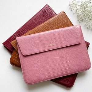 Può includere: Tre custodie rettangolari per computer portatile con texture a stampa coccodrillo, impilate una sull'altra. La custodia superiore è rosa chiaro, quella centrale è color cuoio e quella inferiore è bordeaux scuro. La scritta SIDERA è in oro.