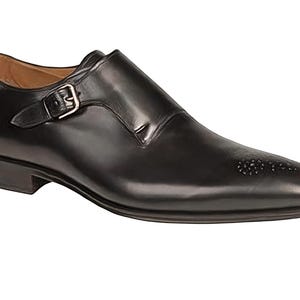 Könnte beinhalten: Ein einzelner schwarzer Lederschuh mit Monk-Strap-Verschluss und silberner Schnalle. Der Schuh hat eine spitze Zehenpartie mit dekorativen Perforationen und einen niedrigen Absatz. Der Schuh ist für formelle Anlässe konzipiert.