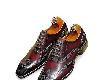 May include: A pair of burgundy leather dress shoes with black toe caps. The shoes feature decorative perforations, gray laces, and a silver ring detail on the heel. The shoes have a polished, glossy finish.