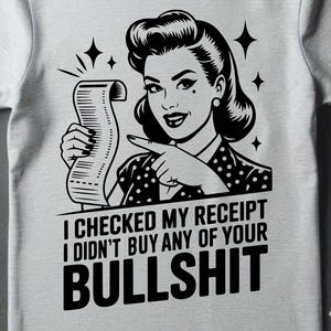 May include: Light gray t-shirt featuring a black and white graphic of a woman holding a receipt and pointing. The text on the shirt reads, "I CHECKED MY RECEIPT I DIDN'T BUY ANY OF YOUR BULLSHIT."