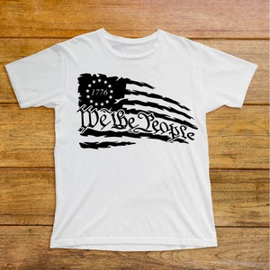 Puede incluir: Camiseta blanca con un dise&ntilde;o de bandera estadounidense desgastada en negro y el texto "We the People" con "1776" en un c&iacute;rculo sobre la bandera.