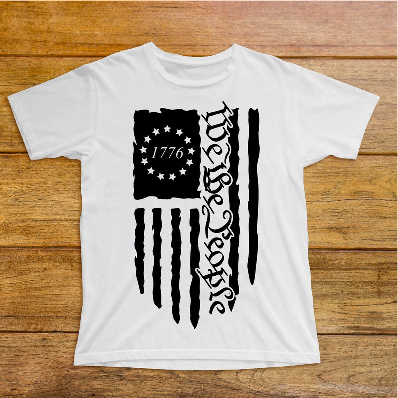 Puede incluir: Camiseta blanca con un gr&aacute;fico en blanco y negro de una bandera estadounidense con el texto "We the People" y "1776" impreso en ella.