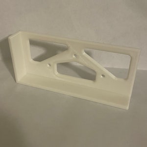 May include: A white, rectangular bracket with a geometric design. The bracket has a flat base and a right-angled corner. The design includes a series of open spaces and three small holes. The object is made of a solid, opaque material.