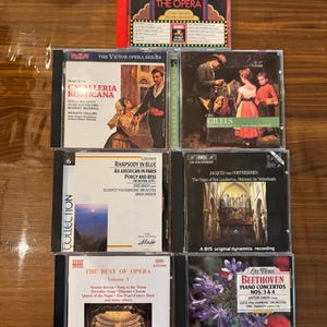 Puede incluir: Una colección de ocho discos compactos de música clásica. Los discos están dispuestos en dos filas de cuatro. La fila superior presenta un disco con un diseño rojo, blanco y azul, un disco con una imagen de una mujer con un vestido rojo, un disco con una imagen de un hombre con un traje negro y un disco con una imagen de un hombre con una camisa blanca. La fila inferior presenta un disco con una imagen de una mujer con un vestido azul, un disco con una imagen de un hombre con un traje negro, un disco con una imagen de un hombre con una camisa blanca y un disco con una imagen de un hombre con un traje negro.