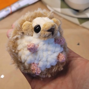 Pode incluir: Um brinquedo de pelúcia de ouriço-cacheiro feito à mão. O ouriço tem um corpo castanho claro e creme com uma textura fofa. Apresenta uma barriga branca, olhos pretos e pés cor-de-rosa de crochê. O brinquedo foi projetado para se assemelhar a um ouriço.