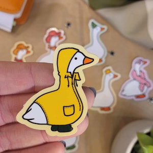 May include: A yellow sticker of a goose in a yellow raincoat with a hood. The goose has a white body, black feet, and an orange beak. Other goose stickers are in the background, some wearing hats and scarves.