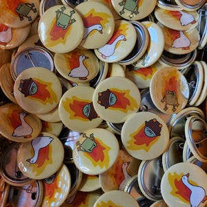 May include: A collection of circular pin-back badges featuring illustrations of a green frog or a white goose surrounded by orange and yellow flames. Each badge has a metal back with a pin mechanism for attachment to fabric or other surfaces.