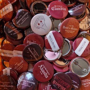 May include: A collection of round pin-back buttons in various shades of red, maroon, and white. The buttons feature printed text including phrases like My coven is Claudia, I have the blood of Akasha in me, and Interview with the Vampire.
