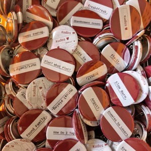 Peut inclure: Une collection de badges ronds avec un design bicolore rouge foncé et blanc. Chaque badge affiche diverses phrases imprimées en noir, telles que Kudos, Hurt/Comfort, Slow Burn, Enemies to Lovers, Found Family et Angst.