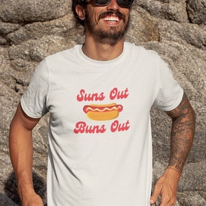 May include: A white t-shirt with a red and yellow hot dog graphic and the text "Suns Out Buns Out". The person is wearing the shirt and standing on a sandy beach.