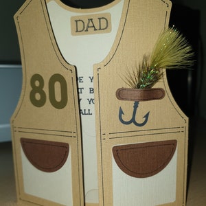 May include: A brown paper card shaped like a fishing vest with the word "DAD" on the front and the number "80" on the left side. The vest has two pockets and a fishing lure on the right side.