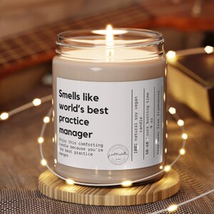 May include: A brown soy wax candle with a white label that reads "Smells like world's best practice manager". The candle is lit and sitting on a wooden coaster.