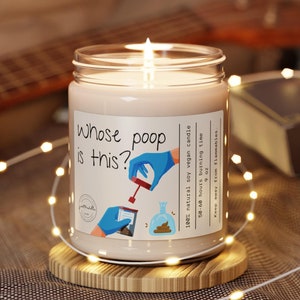 May include: A white soy candle in a clear glass jar with a brown wooden lid. The candle has a label that says "Whose poop is this?" with a cartoon illustration of a hand holding a poop bag. The candle is 100% natural soy vegan and has a burn time of 50-60 hours.