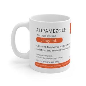 May include: White ceramic mug with a black and orange design. The mug features the text "ATIPAMEZOLE Injectable solution 5 mg/mL Consume to reverse sleepiness and sedation, and to wake your ass up Keep in a safe place. Can also be used as a pen holder. For veterinary use only Rapid onset of reversed sleepiness for veterinary"