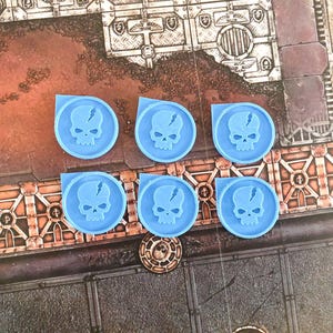 Könnte beinhalten: Sechs blaue Plastik-Schädelmarker mit einem Riss im Schädel. Die Marker sind rund und haben ein weißes Schädelmotiv.