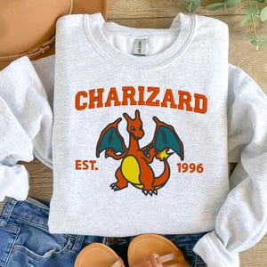 May include: Gray sweatshirt with a red, orange, and yellow cartoon of Charizard, a Pokemon character, with the text "CHARIZARD EST. 1996" printed on the front.