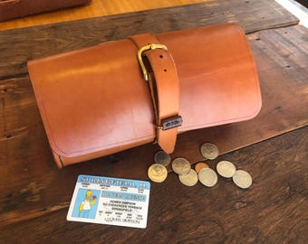 May include: A brown leather travel case with a brass buckle and strap. The case is closed and lying on a wooden surface. There are several coins scattered around the case.