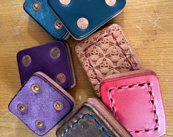 May include: A collection of handmade leather dice boxes in various colors, including teal, purple, red, and brown. Each box is square with rounded corners and features copper rivets. One box has a floral embossed design.