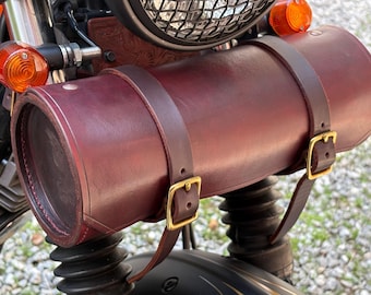 May include: A burgundy leather motorcycle tool bag, cylindrical in shape, mounted on the front of a motorcycle. The bag is secured with brown leather straps and gold-colored buckles. The motorcycle has orange turn signals and a black front end.