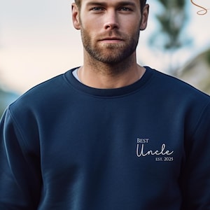 Può includere: Felpa blu navy con la scritta "Best Uncle Est. 2025" in corsivo bianco. La felpa è a tinta unita e ha lo scollo tondo. L'immagine include anche una campionatura di colore che dice "Navy".