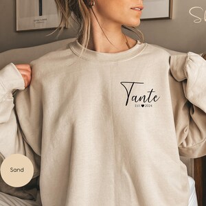 May include: A sand-colored sweatshirt with the text "Tante Est 2024" printed on the front.