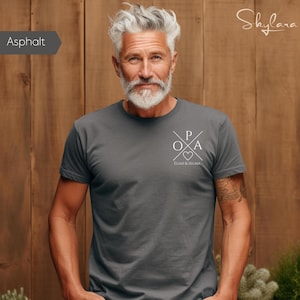 Puede incluir: Camiseta gris asfalto con un diseño gráfico blanco. El diseño incluye líneas cruzadas, las letras P, O y A, un corazón y los nombres Elias & Selma. La camiseta es de manga corta y está hecha de un material suave.