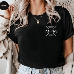 May include: Black t-shirt with a white graphic design that says "MOM" with a heart in the middle. The graphic also includes the text "Constantin Est. 2021" and "Gabriella Est. 2024".