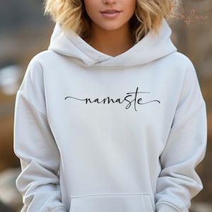 K&ouml;nnte beinhalten: Wei&szlig;er Kapuzenpullover mit dem Wort "namaste" in schwarzer Kursivschrift.