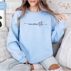 K&ouml;nnte beinhalten: Ein hellblauer Pullover mit dem Wort "namaste" in schwarzer Kursivschrift.