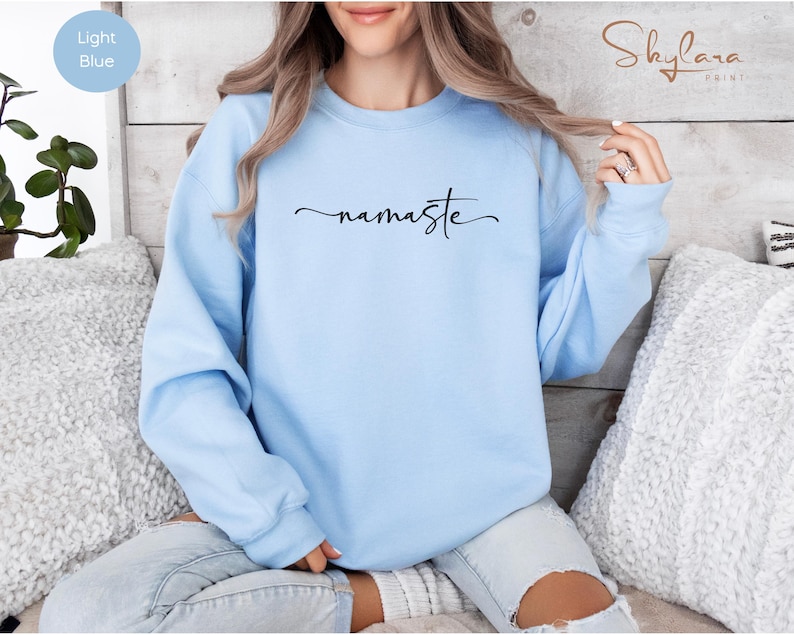 K&ouml;nnte beinhalten: Ein hellblauer Pullover mit dem Wort "namaste" in schwarzer Kursivschrift.