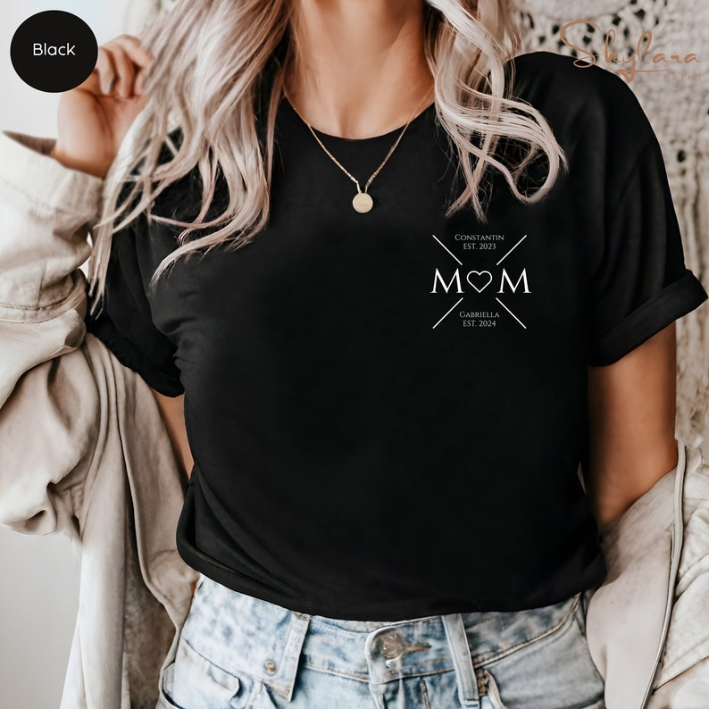 May include: Black t-shirt with a white graphic design that says "MOM" with a heart in the middle. The graphic also includes the text "Constantin Est. 2021" and "Gabriella Est. 2024".